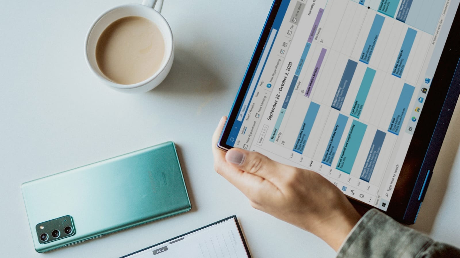Main tenant une tablette affichant un agenda hebdomadaire, posée près d'un téléphone et d'une tasse
