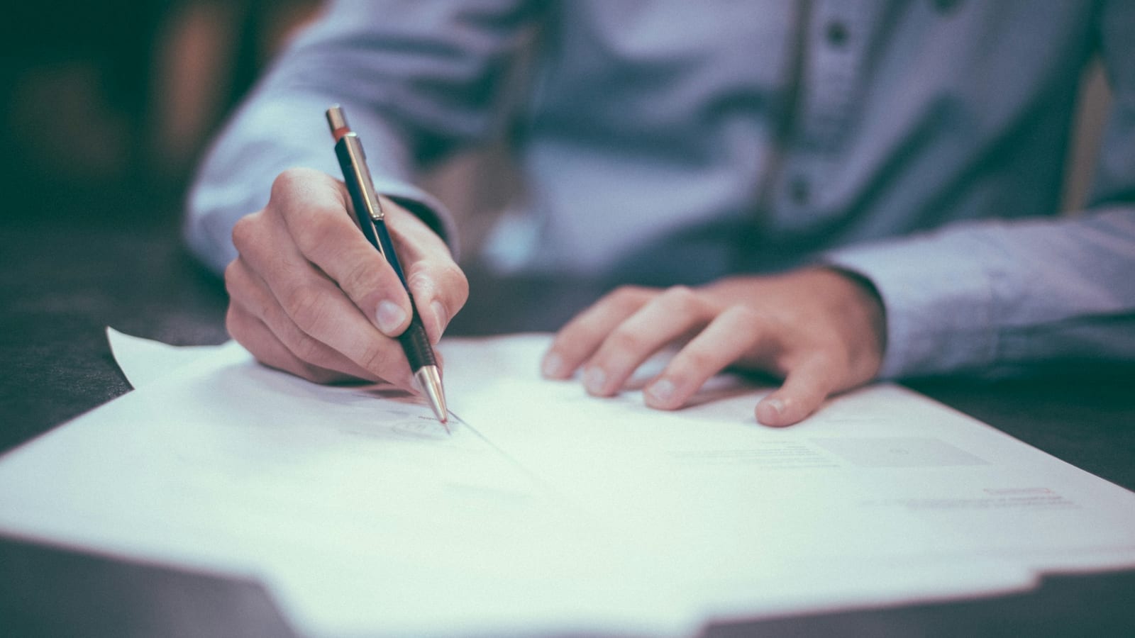 Personne signant un document contractuel, stylo en main, sur un bureau sombre
