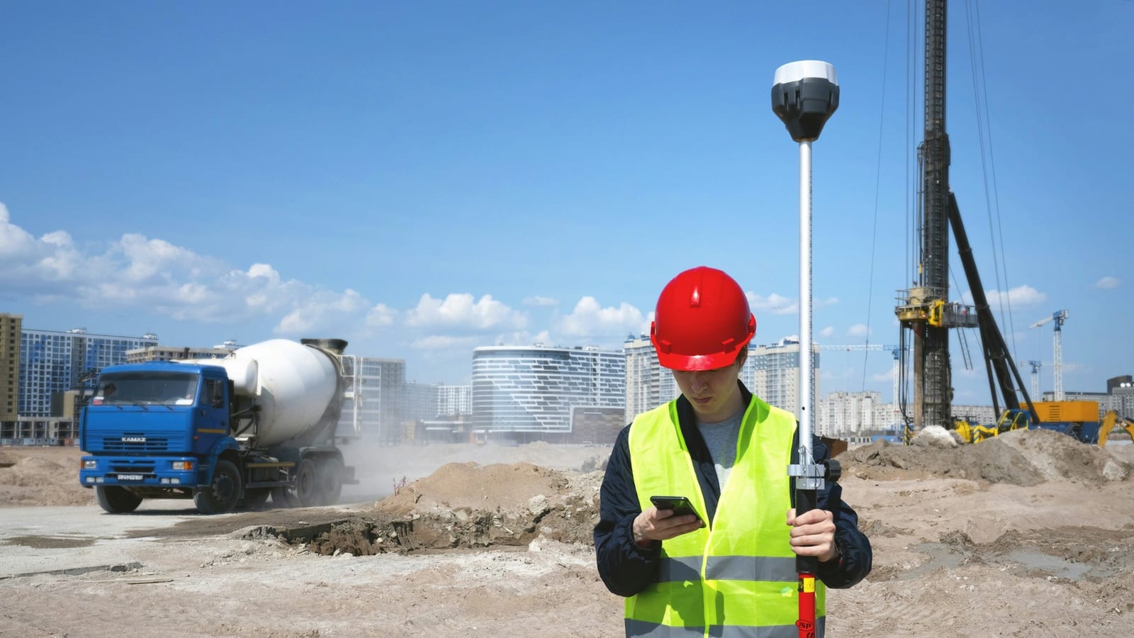 Ouvrier en gilet haute visibilité et casque rouge consultant son téléphone sur un chantier, canne GNSS à la main