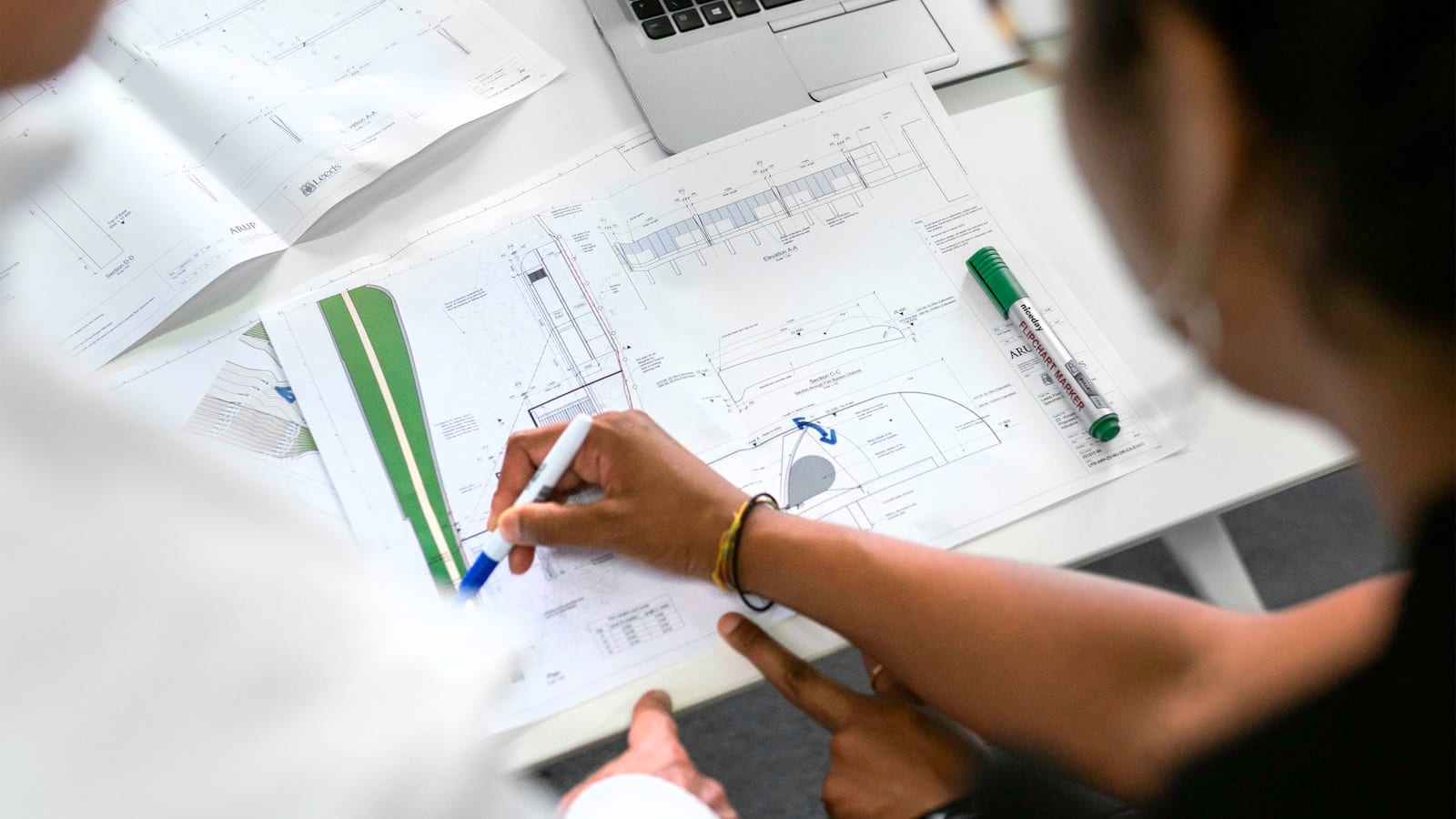 Deux personnes analysant des plans de projet sur un bureau, avec un ordinateur portable